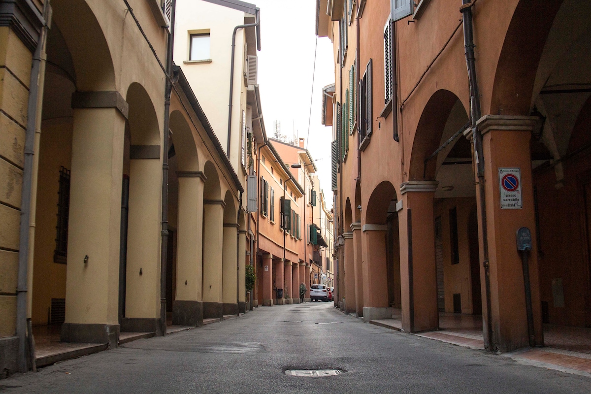 A quiet street in Bologna features arched porticoes along both sides, providing shade and character. The narrow roadway is lined with pastel-colored buildings, showcasing traditional architecture. A parked car is visible in the distance, and cobblestones can be seen under the soft lighting.