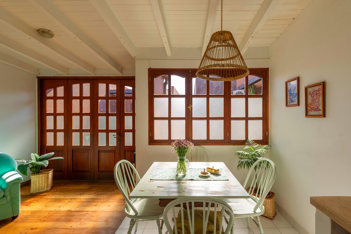 A dining area is visible, featuring a light-colored table surrounded by six chairs. A rattan pendant light hangs overhead, and a bouquet of flowers is placed on the table. Large windows with wooden frames provide natural light, illuminating the wooden floor and green accents in the room.