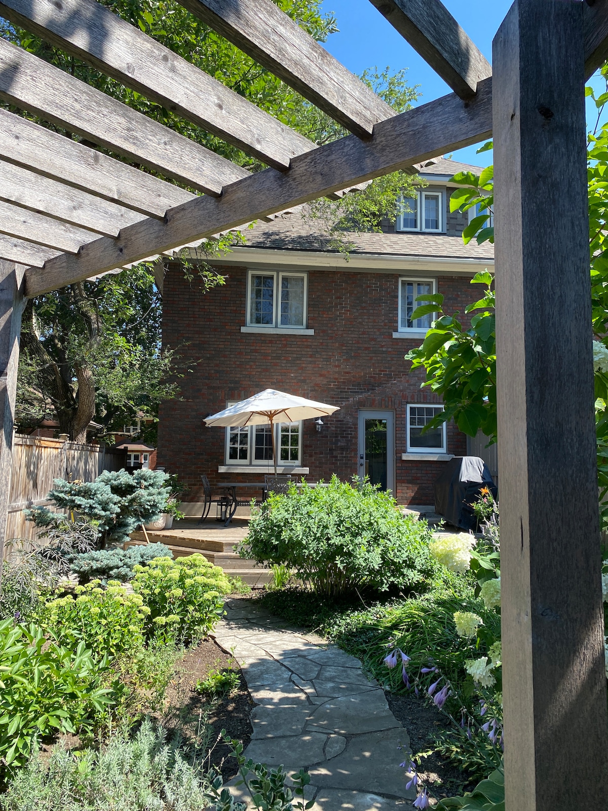 The rear of the home is framed by a wooden pergola, leading to a lush garden filled with varied greenery and blooming flowers. A stone pathway guides to a patio area with a table and umbrella, ideal for outdoor relaxation.