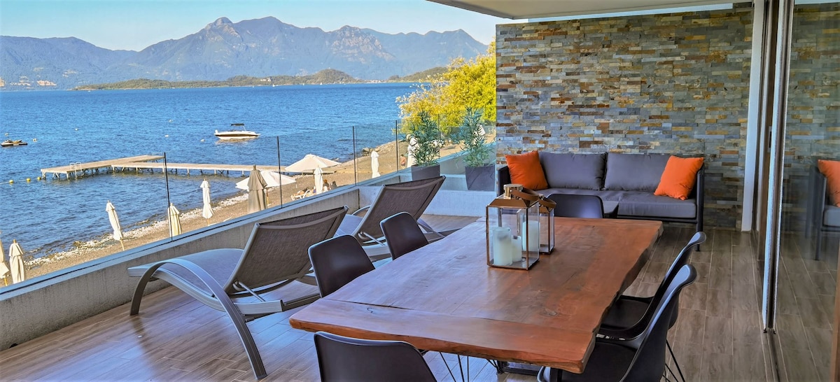 An outdoor terrace is shown with a natural wood dining table surrounded by black chairs. Comfortable lounge chairs are positioned nearby, offering views of the lake and mountains. Large sliding doors frame the space, enhancing the connection with the scenic environment.