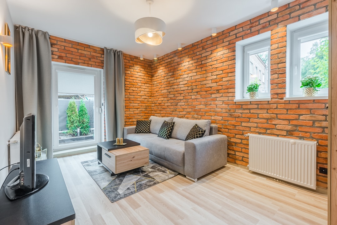 A cozy living area features a textured brick wall that adds character. Light-colored flooring complements a spacious gray sofa adorned with patterned cushions. Two windows provide natural light, while a minimalist coffee table sits atop a patterned rug. A television is positioned on a low wooden unit.