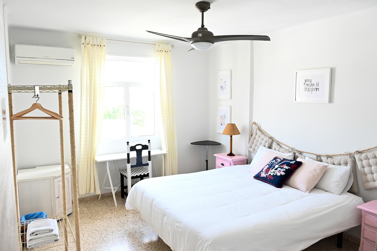 A bright bedroom features a spacious bed framed by light-colored walls. A ceiling fan hangs above, with air conditioning mounted on the wall. Nearby, a small study area is set up with a desk and chair next to a window, allowing natural light to enter.