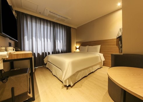 A well-lit guest room is presented, featuring a spacious bed with a neutral-colored coverlet. Natural light filters through sheer curtains, illuminating the minimalist decor, which includes a small wooden desk and a round table. Soft tones are utilized throughout, creating a calm environment.