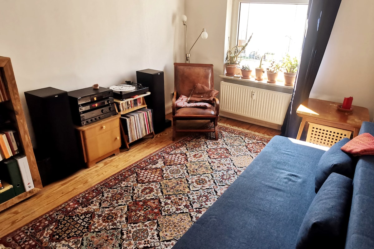 A cozy living space features a dark blue sofa across from a vintage leather chair. A wooden side table sits nearby, alongside speakers and a record player on a low console. Plants in pots are arranged on the windowsill, and a patterned rug covers the wooden floor.