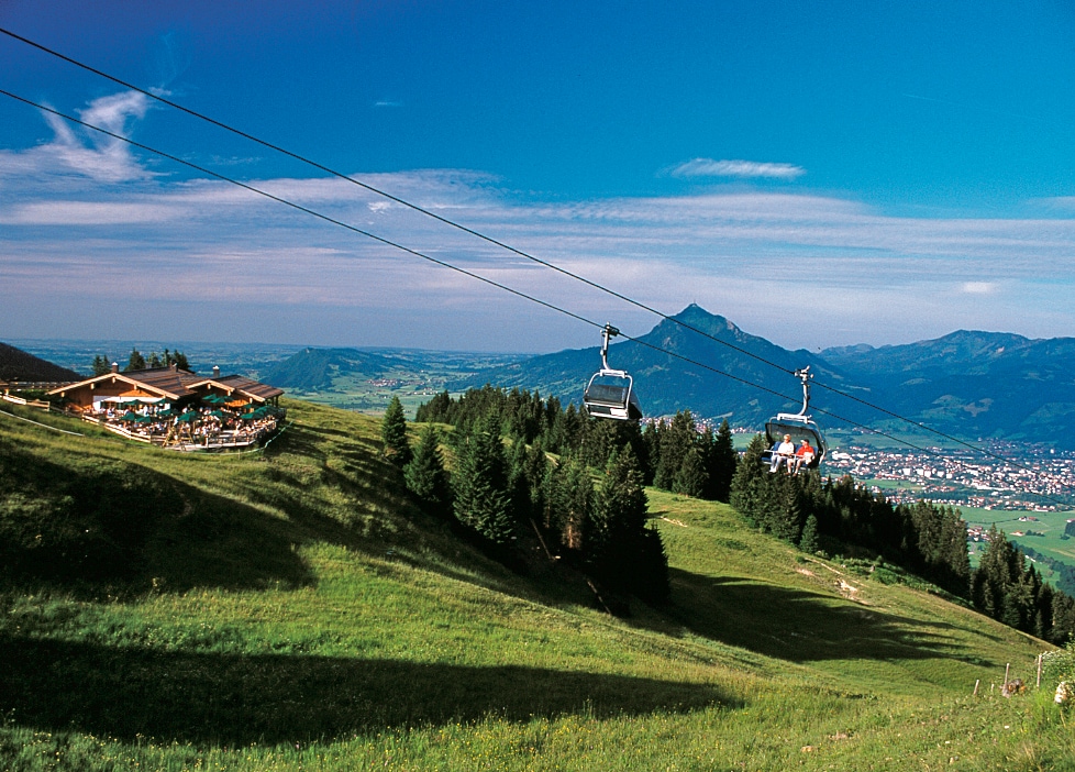 A scenic landscape displays a ski lift transporting visitors over lush green hills, with a restaurant nestled among the trees below. The backdrop features distant mountains under a clear blue sky, creating a relaxing and natural environment.