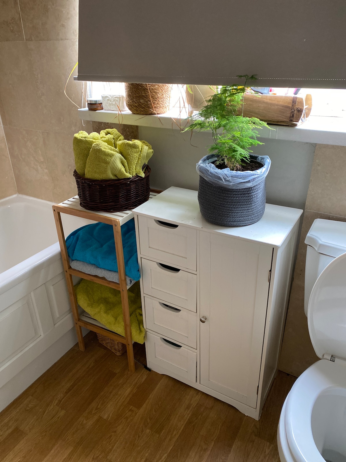 A bathroom corner features a white cabinet with drawers, alongside a bamboo shelf holding colorful towels in blue and green. A small potted plant is placed on the shelf, adding a touch of greenery, while natural light enters through a window dressed with a simple blind.