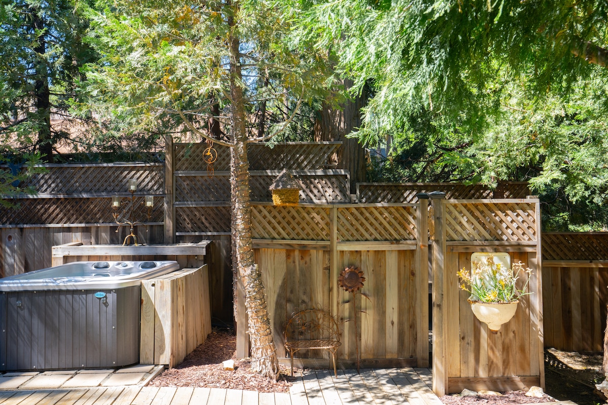 The outdoor space features a private jacuzzi surrounded by wooden fencing and greenery. A small decorative wall-mounted planter is visible along with a wooden bench. The area is designed for privacy, with natural elements integrated into the setting.