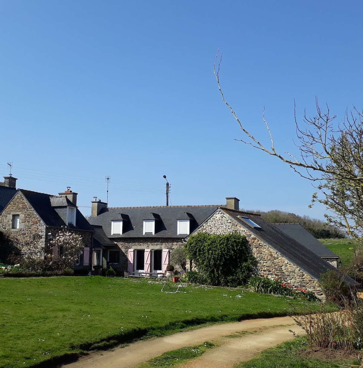 The exterior view displays a charming stone house with a slate roof, surrounded by a lush green garden. A curved pathway leads to the entrance, and bright blue skies provide an inviting backdrop to the scene.