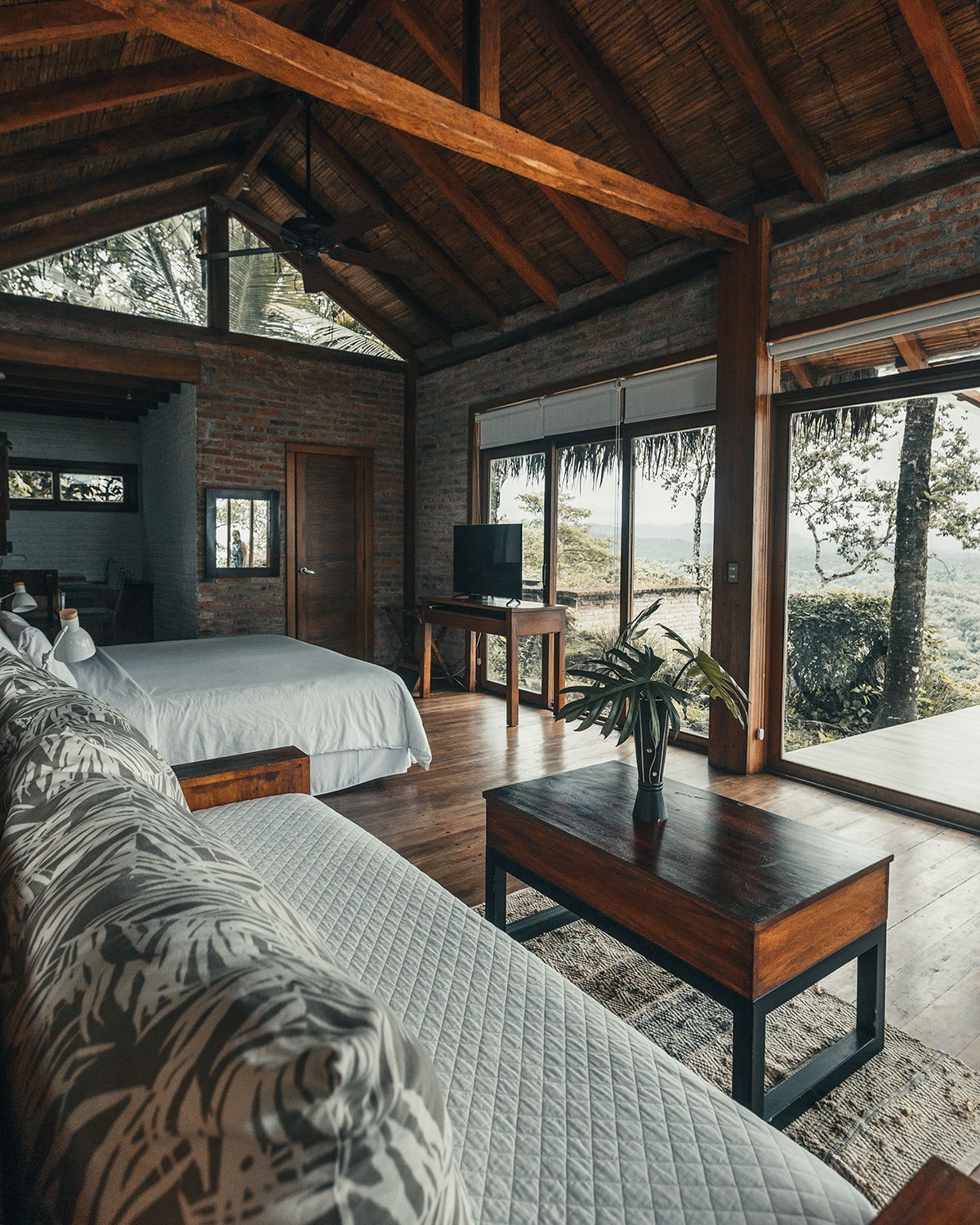 A spacious interior area features high wooden ceilings and large windows that provide views of the surrounding nature. A cozy bed is positioned next to a wooden table and a comfortable sofa, creating a welcoming and relaxing atmosphere.