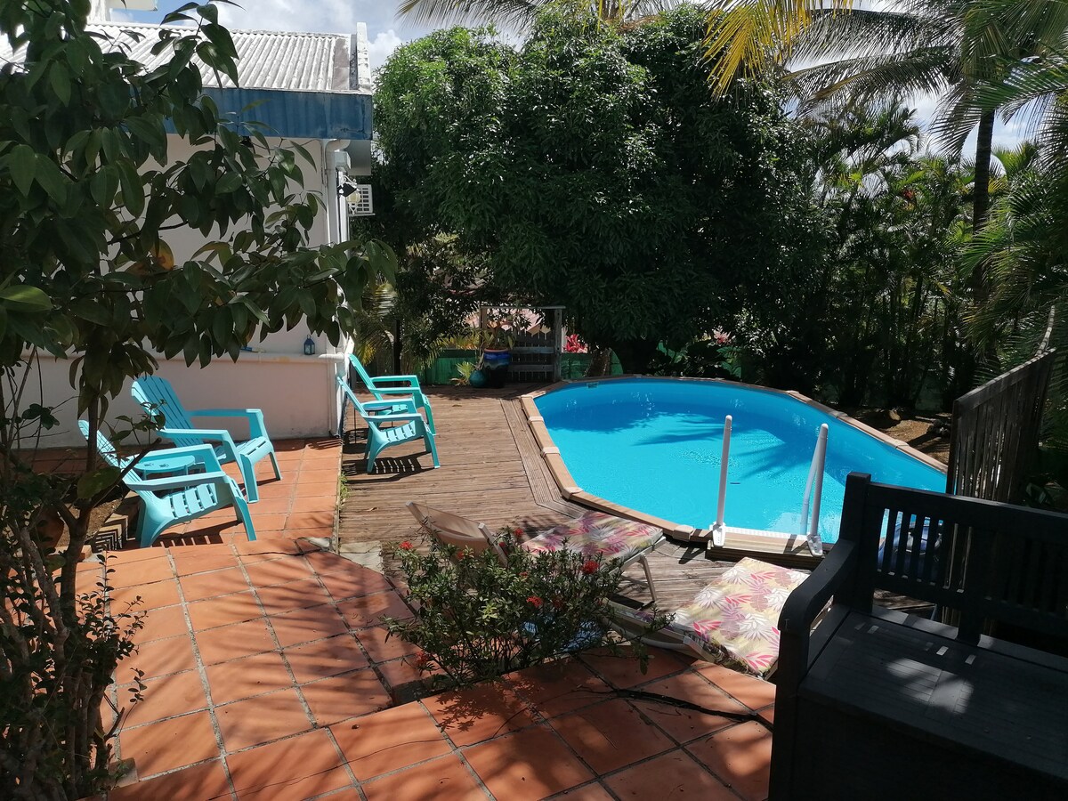 A private pool is surrounded by a wooden deck and colorful lounge chairs. Lush greenery enhances the setting, providing privacy. The pool features easy access steps, and a relaxing atmosphere is created by the combination of sun and shade from nearby trees.
