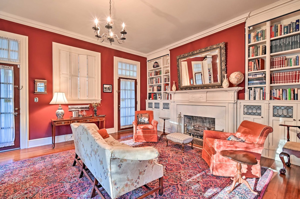 A cozy living room with warm red walls is highlighted by a central fireplace and built-in bookshelves. Comfortable seating includes an upholstered sofa and armchairs. Natural light enters through large windows, enhancing the inviting atmosphere. Decorative elements include a chandelier and wall art.