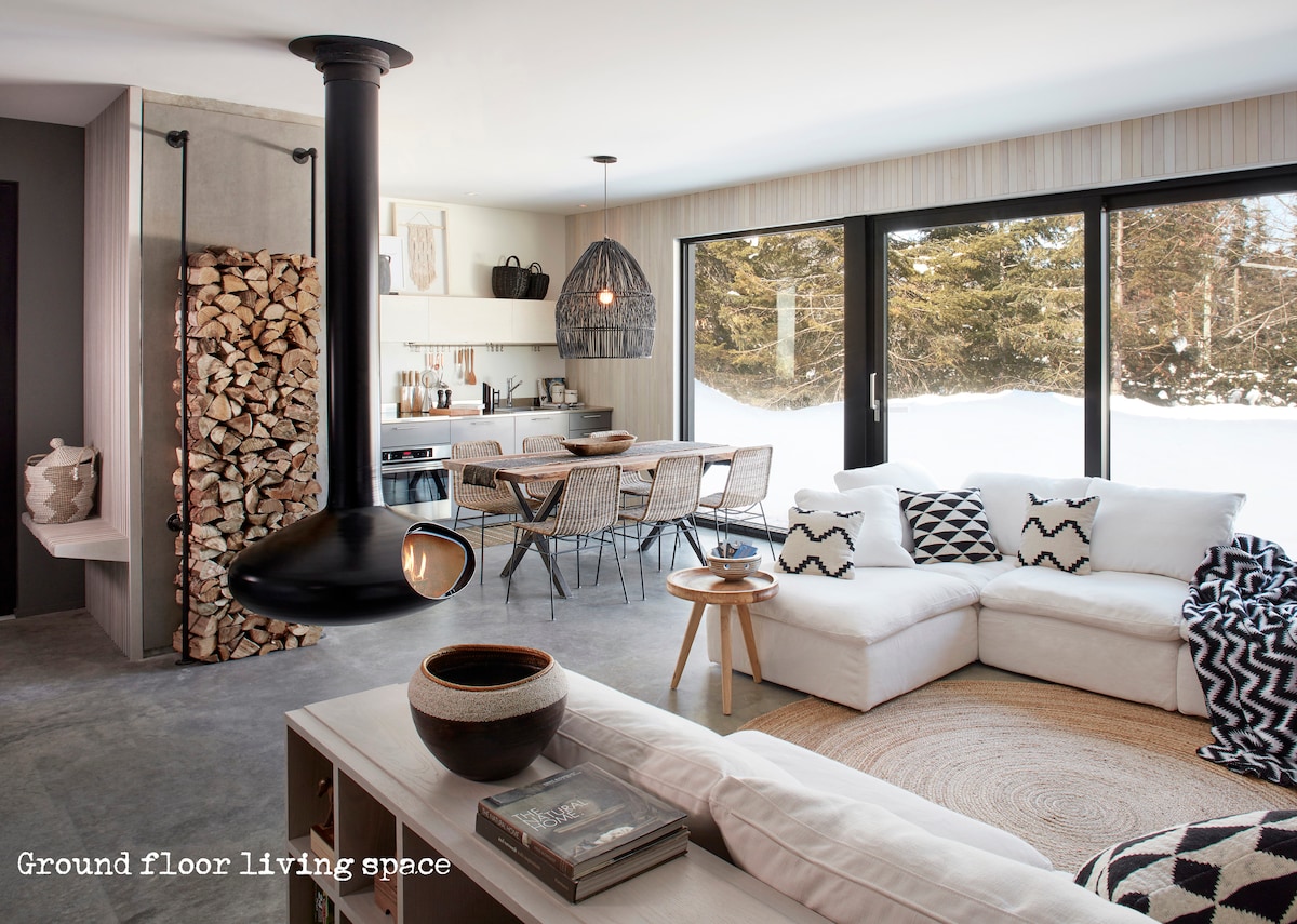 The ground floor living space features a modern white sectional sofa accented with black and patterned cushions. A suspended black fireplace enhances warmth, while a wooden table with woven chairs is positioned in the background. Large windows offer views of the snow-covered outdoor landscape.
