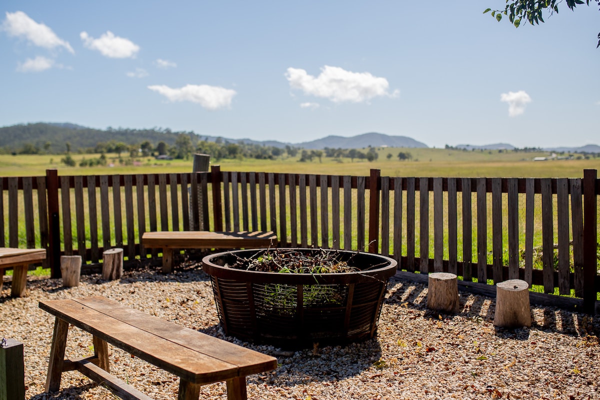 An open outdoor fire pit area is surrounded by wooden benches and tree stump seating. A wooden fence encloses the space, offering views of lush farmland and rolling hills under a clear blue sky. The setting invites relaxation and outdoor gatherings.