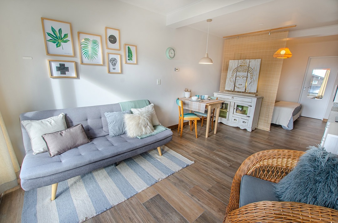 A cozy living area is presented with a gray sofa bed adorned with textured cushions, complemented by a woven chair. A light wood dining table and blue chairs are visible, along with framed botanical art on the walls, creating a relaxed ambiance.