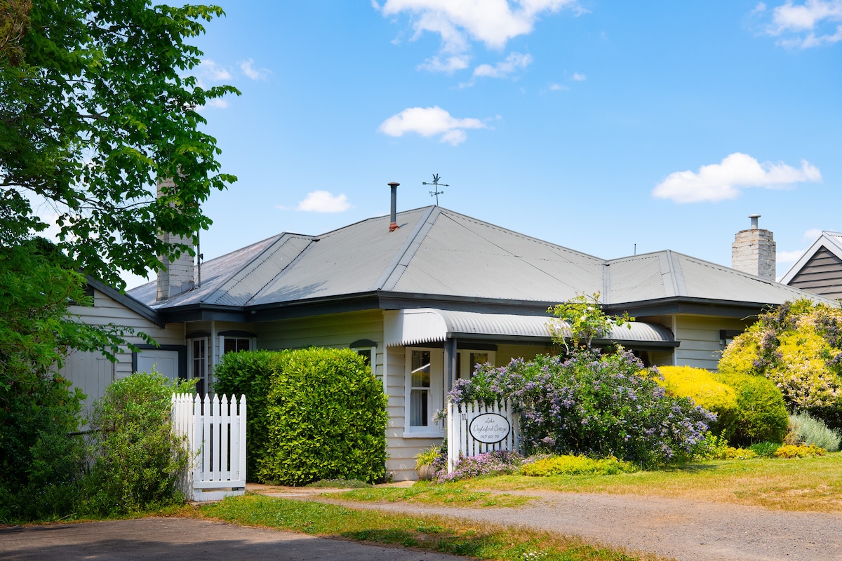 The exterior of a charming cottage is framed by lush greenery and colorful blooms. A white picket fence fronts the property, leading to a welcoming entrance. The sloped roof, traditional architecture, and well-maintained garden create an inviting atmosphere near Lake Daylesford.