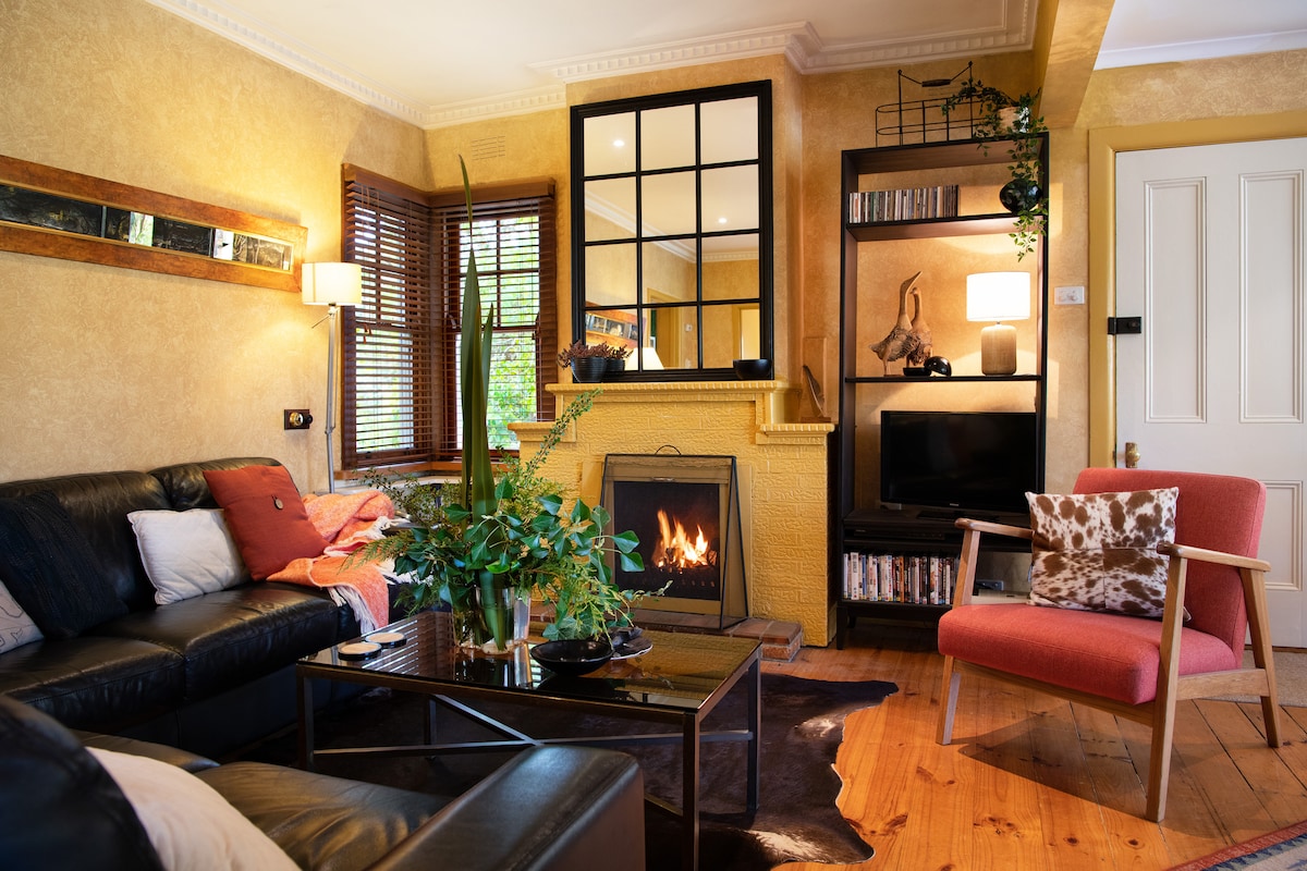 An intimate living area is showcased with a fireplace and large mirror. A leather sofa and a red armchair provide seating. Shelves display books and decor, while natural light filters through the window, illuminating the warm walls and plant accents.