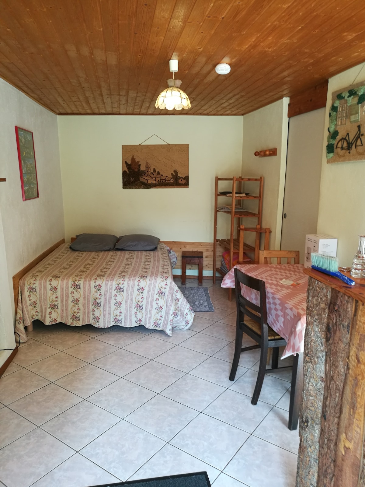 A cozy studio is displayed with a spacious bed covered in a floral duvet, complemented by soft pillows. A small wooden dining table with two chairs is positioned near a shelf. Light is provided by a hanging fixture, and a textured wall adds warmth to the space.