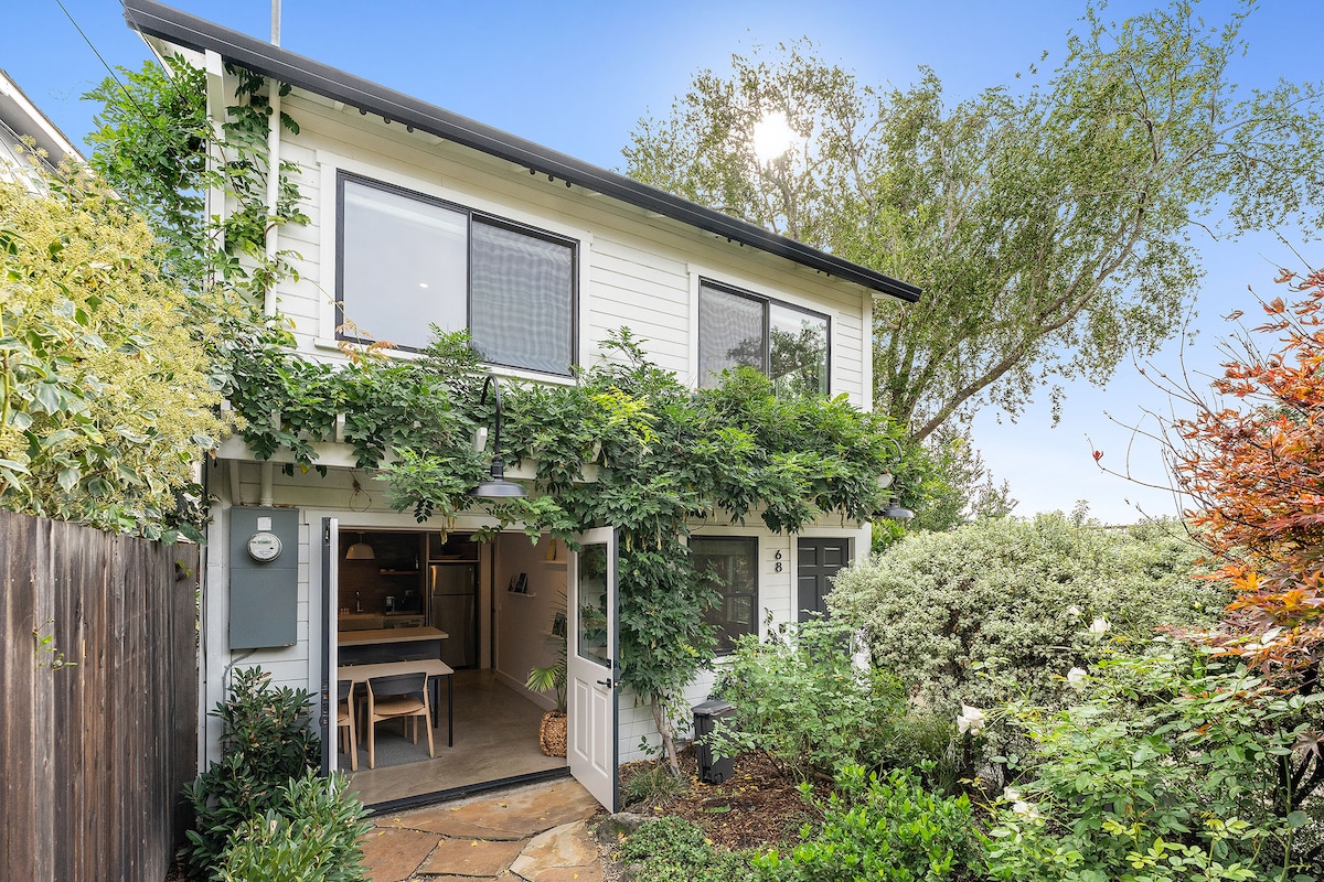 The exterior of the casita is surrounded by lush greenery, with a charming Dutch door opening into the space. Sunlight filters through the leaves, highlighting the inviting entrance and the well-maintained garden area that adds to the serene ambiance.