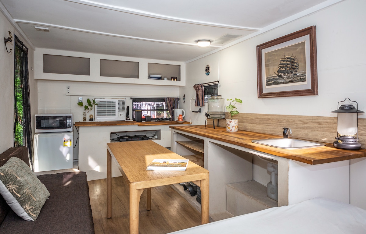 An interior view of a renovated caravan showcases a streamlined kitchen area with essential appliances, including a microwave and a small refrigerator. A wooden table serves as a dining or workspace, accompanied by a comfortable seating area. Decorative plants add a touch of greenery to the space.