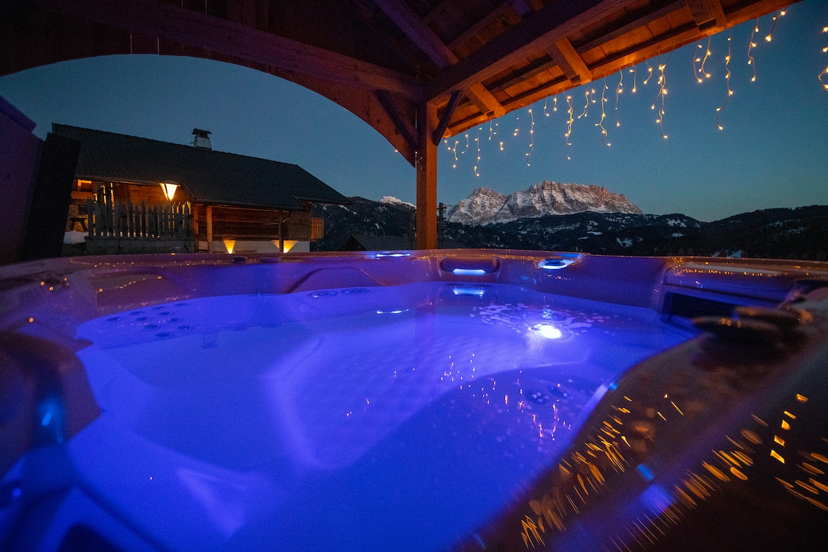 An outdoor hot tub is illuminated with soft blue light, situated under a rustic wooden shelter. Views of majestic mountain peaks are visible in the background, while twinkling fairy lights adorn the structure, creating a serene ambiance for relaxation.
