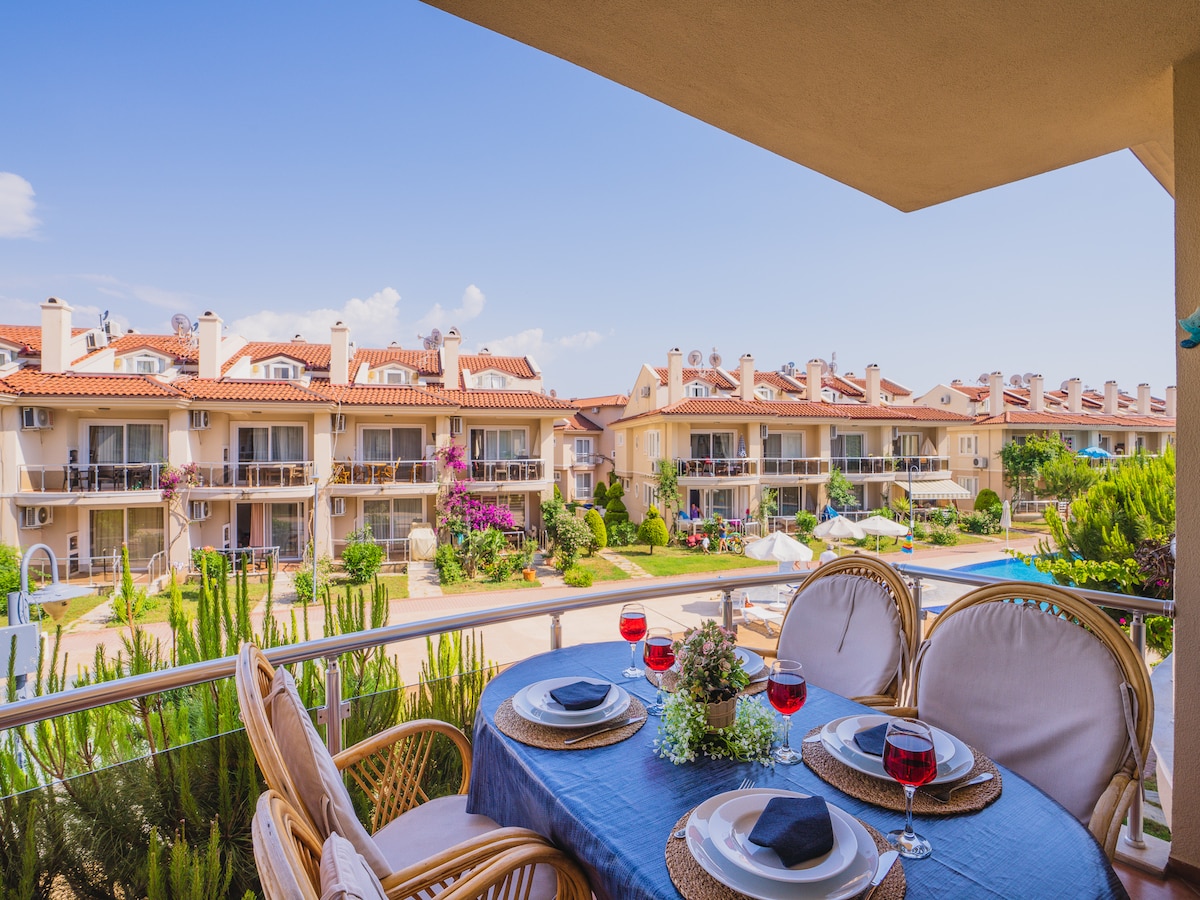 An outdoor dining area is set up on a balcony, featuring a table with a blue tablecloth and four plates, complemented by glasses of red drink. Lush greenery surrounds the space, while the view captures several villa-style buildings with terracotta roofs in the background.