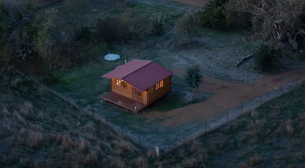 A cozy wooden cabin with a red roof is surrounded by open paddocks and trees. Warm light glows from its windows, highlighting the inviting exterior. The cabin is set on a grassy area, bordered by a wire fence, with a water tank visible in the background.