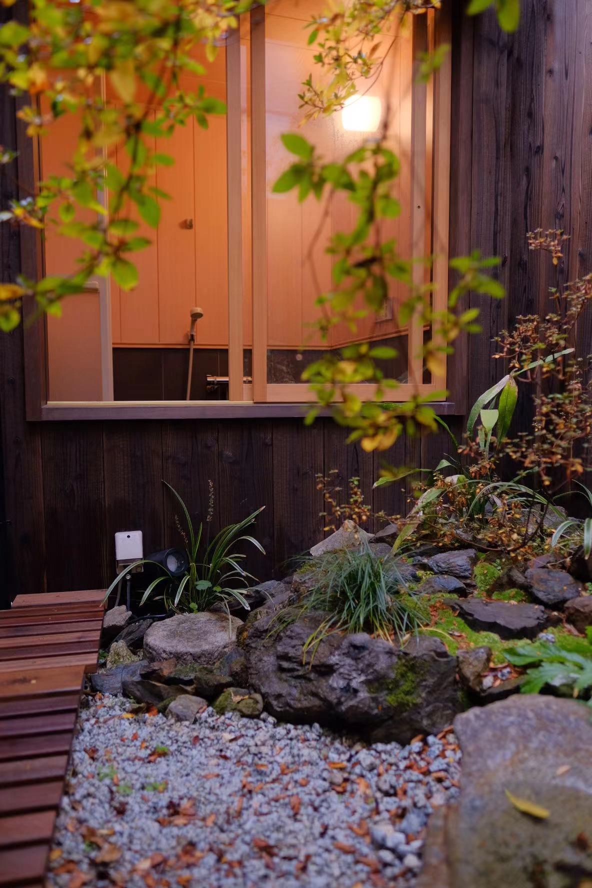 A tranquil private garden is highlighted, featuring carefully arranged rocks and green plants. A wooden pathway leads towards a window that hints at a bathing area inside. Soft lighting illuminates the space, creating a serene ambiance.