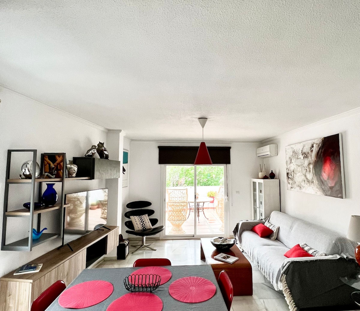 The living area features a modern design with a grey sofa and a black armchair complemented by a vibrant artwork. A dining table with red accents is visible, alongside minimalist shelves displaying decorative items. Large glass doors open to a terrace surrounded by greenery.