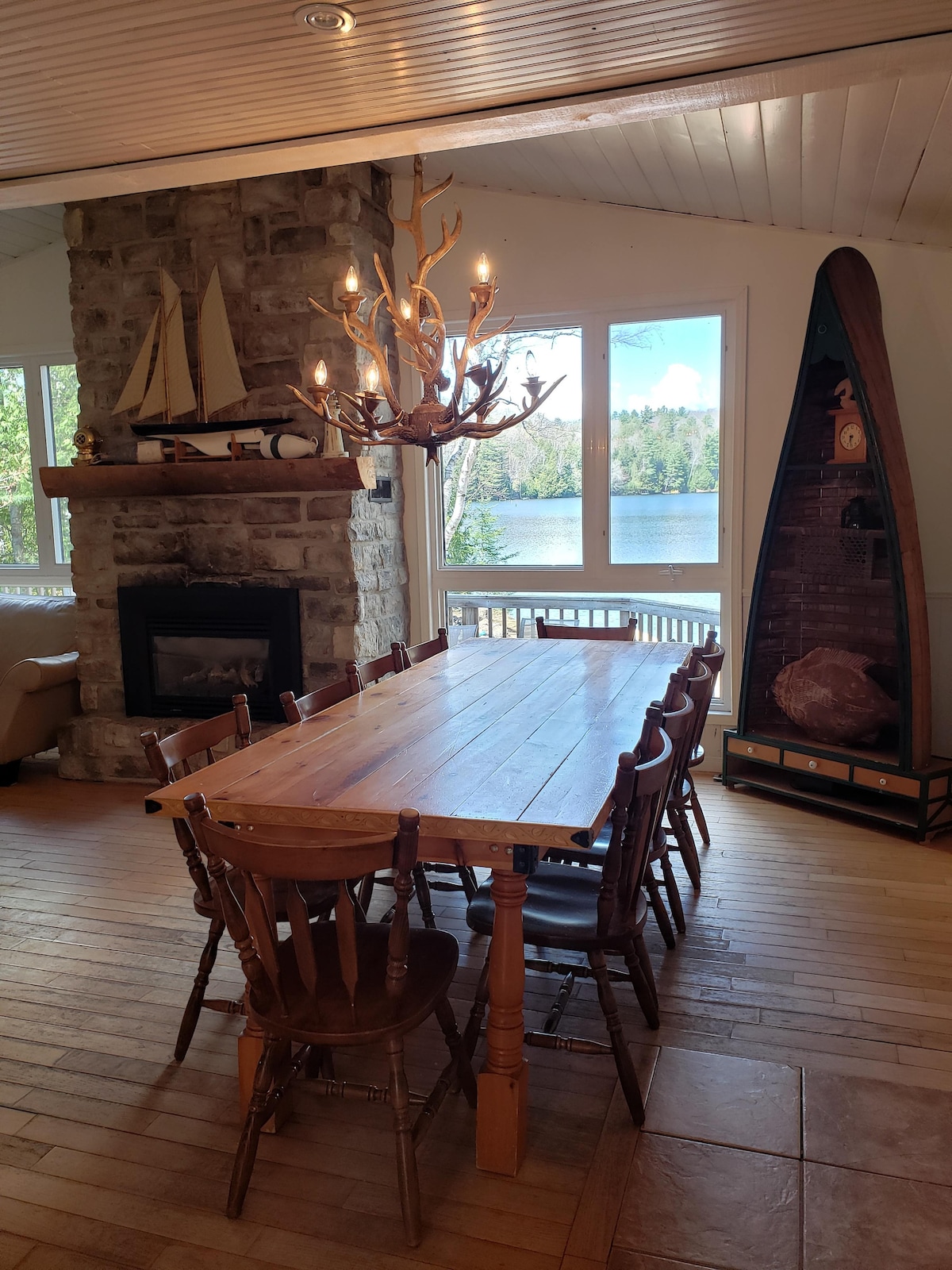 An open-concept dining area features a large wooden table surrounded by eight wooden chairs. Natural light enters through multiple windows, revealing views of the lake outside. A stone fireplace and decorative elements, including a canoe, add a rustic charm to the space.