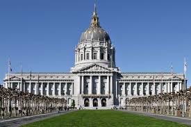 The image showcases the grand exterior of City Hall, characterized by its iconic dome and ornate architectural details. Flanking paths lead through well-manicured green lawns, creating a sense of openness. The clear blue sky provides a vivid backdrop, emphasizing the building's prominent stature.