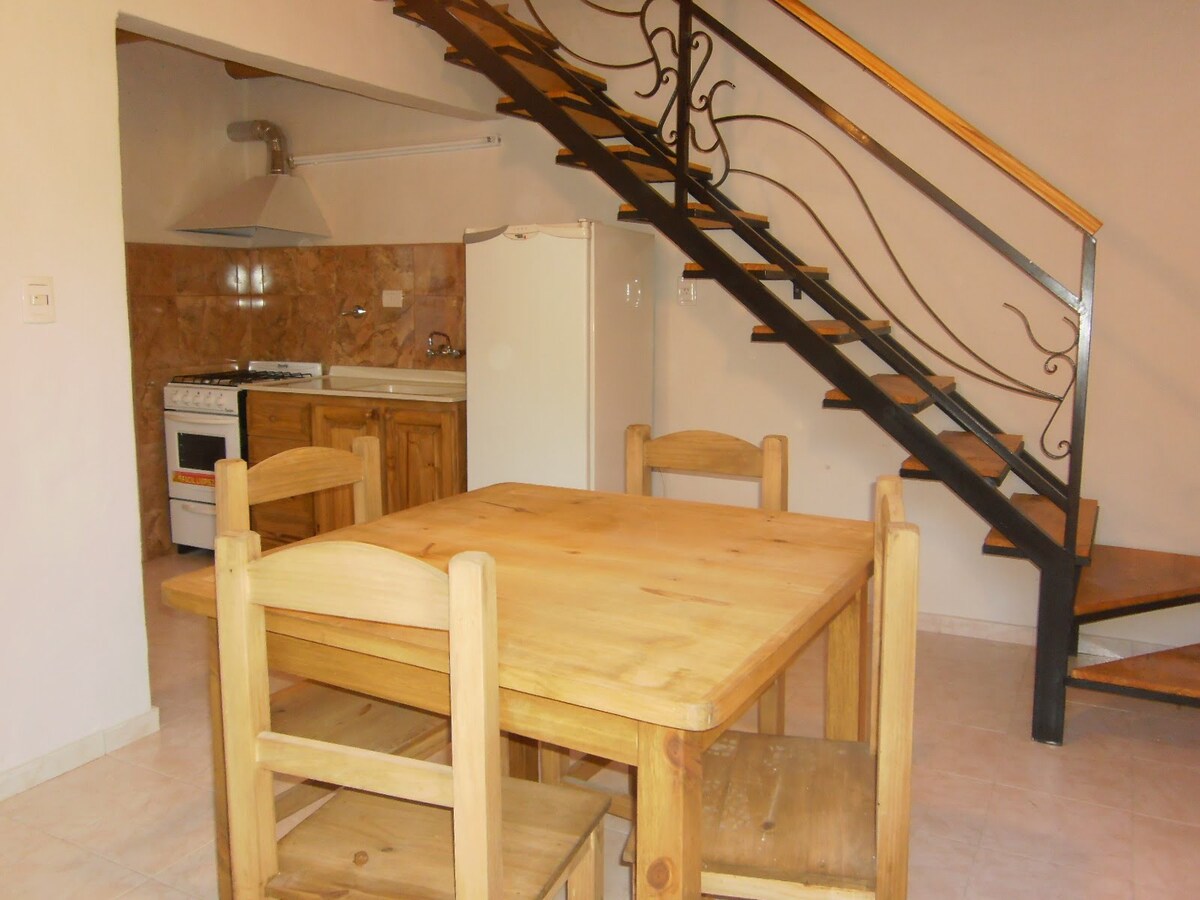 A wooden dining table with four matching chairs occupies the center of the room. A staircase with decorative iron railings leads to the upper level. In the background, a kitchenette is visible, featuring a gas stove, refrigerator, and wooden cabinetry against a tiled backsplash.