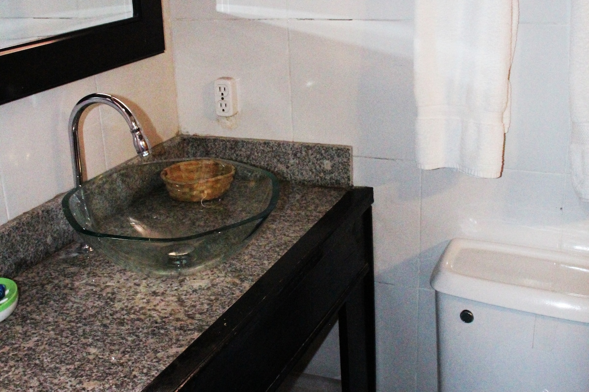A modern bathroom is featured with a stylish glass sink set atop a granite counter. An elegant bowl is placed beside the sink. A white toilet is positioned against the wall, complemented by white towels hanging on a nearby rack.