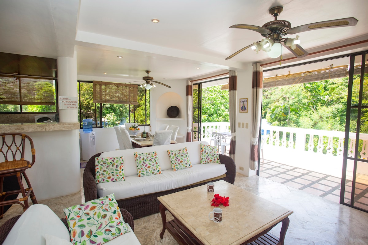 A comfortable living area features a white sofa adorned with floral cushions, complemented by a light-colored coffee table. Large glass doors open to a terrace garden, allowing ample natural light. Ceiling fans above provide a cooling breeze, enhancing the relaxed ambiance.