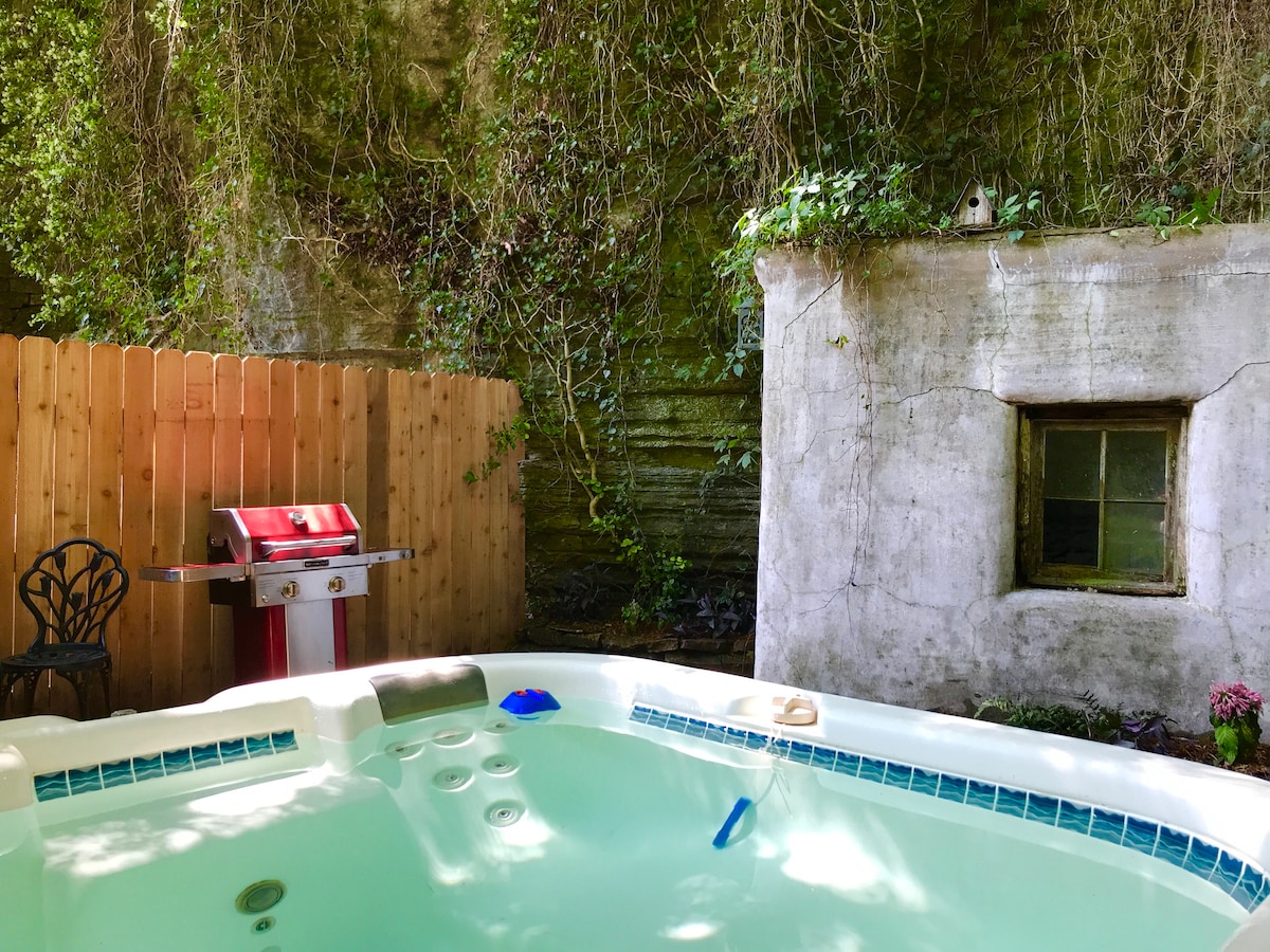 A private hot tub is featured in a secluded outdoor area, surrounded by lush greenery and a wooden fence. A charcoal grill is positioned nearby on a stone pathway, with a vintage-style window visible in the rustic wall.