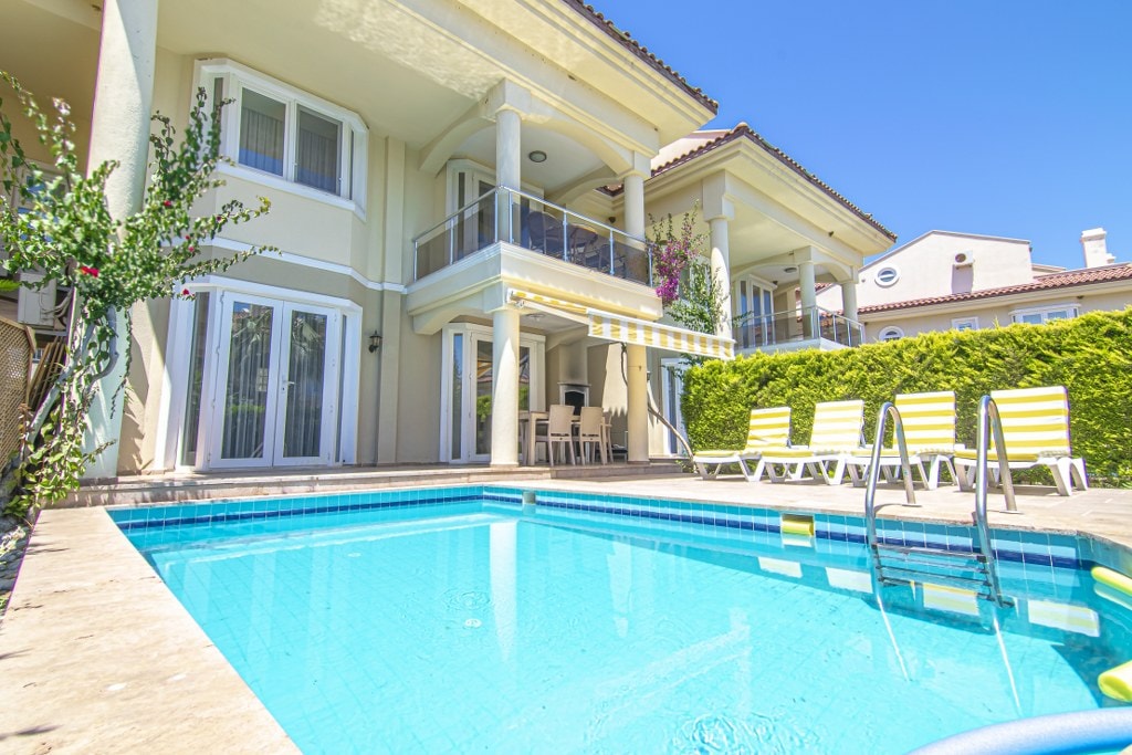 A private pool is framed by a stone patio, with yellow and white lounge chairs positioned nearby. The exterior of a two-story villa is visible, featuring large glass doors and a balcony adorned with greenery, creating an inviting outdoor space.