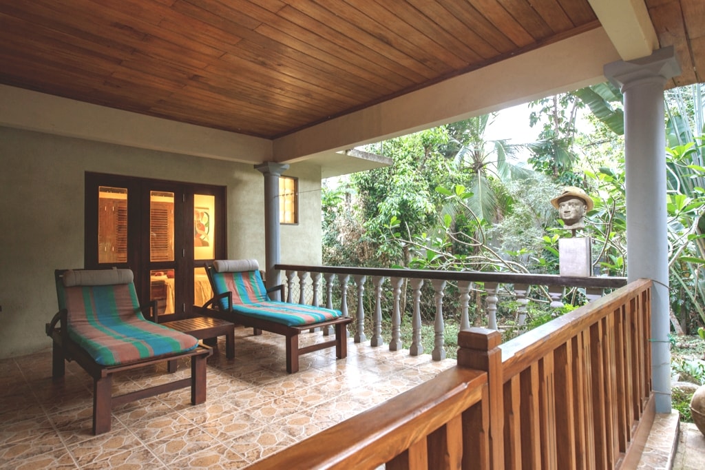 A spacious verandah features two lounge chairs with colorful cushions, inviting relaxation amidst a lush garden backdrop. The wooden railings surround the area, and large glass doors lead into the living space, allowing natural light to enter.