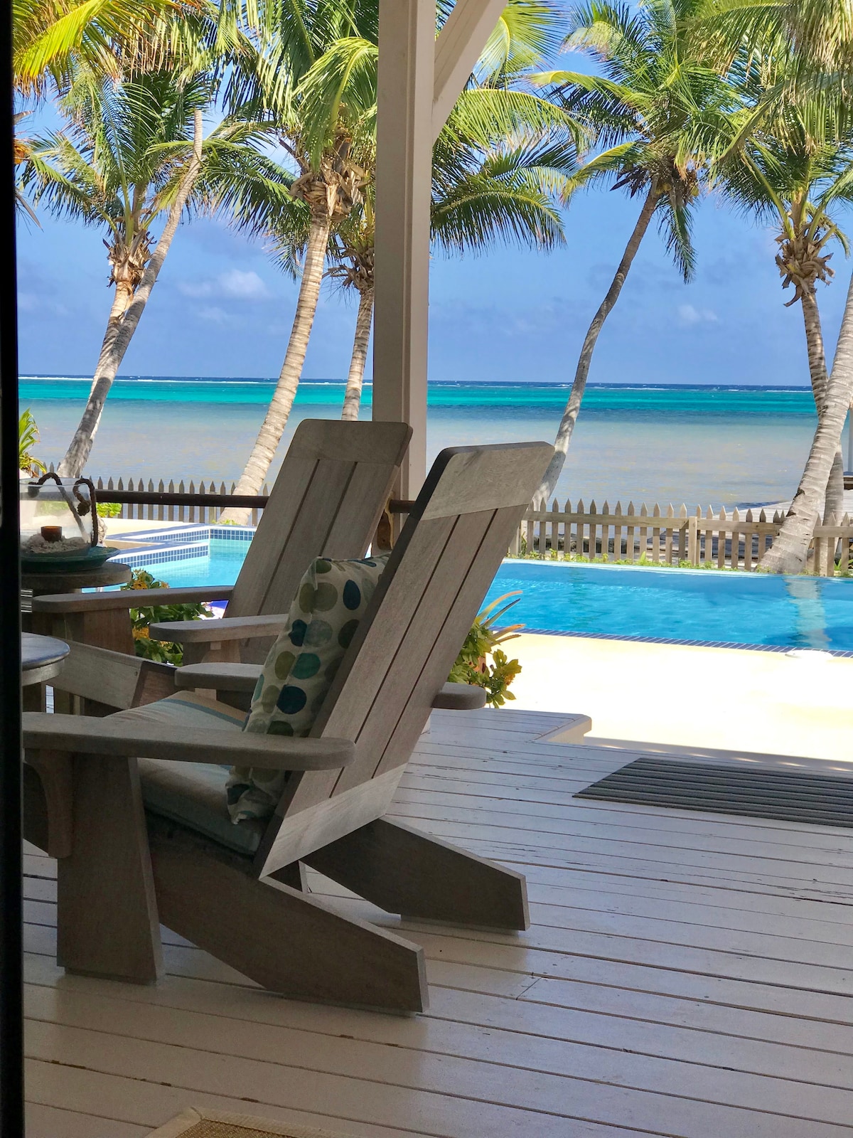 Two comfortable wooden adirondack chairs are positioned on a covered deck, overlooking a tranquil pool and the Caribbean Sea. Lush palm trees frame the view, with a bright blue sky providing a serene backdrop.