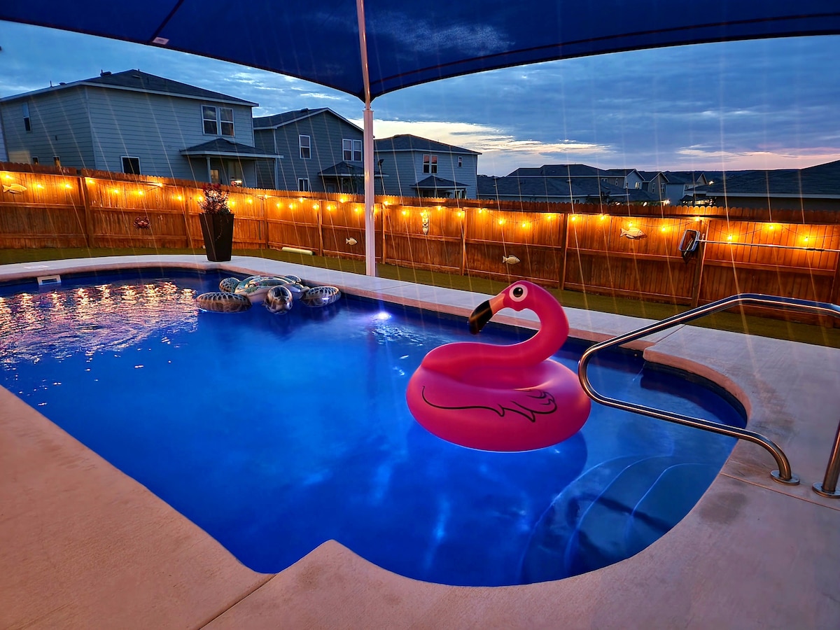 A clear swimming pool is surrounded by a spacious patio and a boundary fence. A pink flamingo float rests on the water, while string lights provide a soft glow above the patio area. The evening sky creates a serene backdrop.
