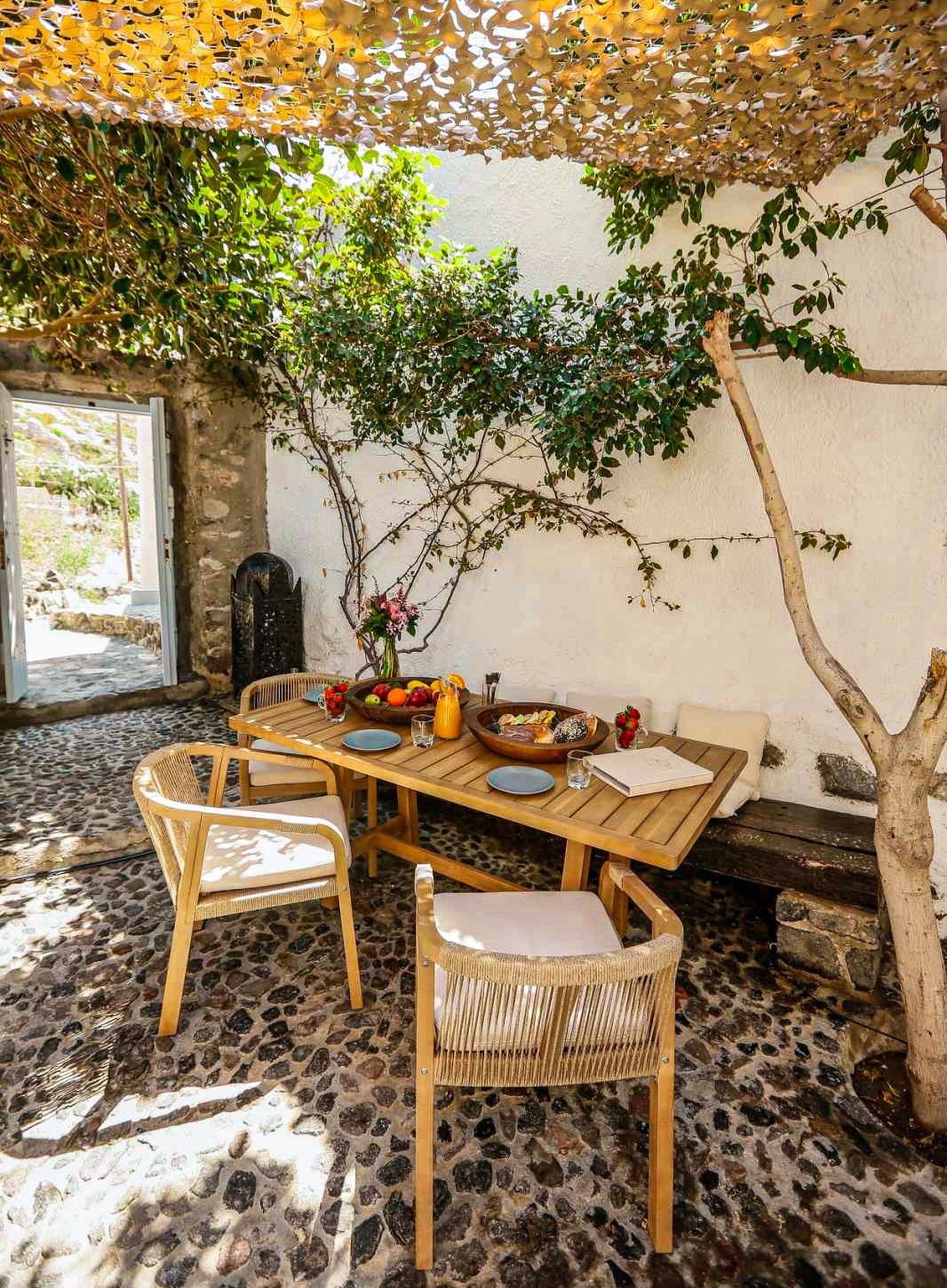 In a charming courtyard setting, a wooden dining table is surrounded by four light-colored chairs. The table is elegantly set with fresh fruits and drinks, while lush greenery and a stone wall create a serene atmosphere. Notable pebbled flooring enhances the rustic charm.