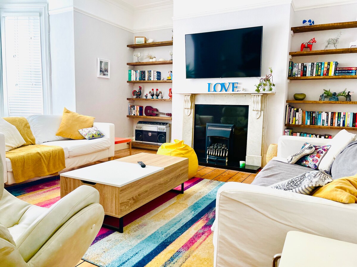 A bright living room is featured, equipped with comfortable seating including white sofas and a stylish armchair. A colorful striped rug adds vibrancy. Shelves with books and decorative items line the walls. A flat-screen TV is mounted above a decorative fireplace, enhancing the cozy environment.