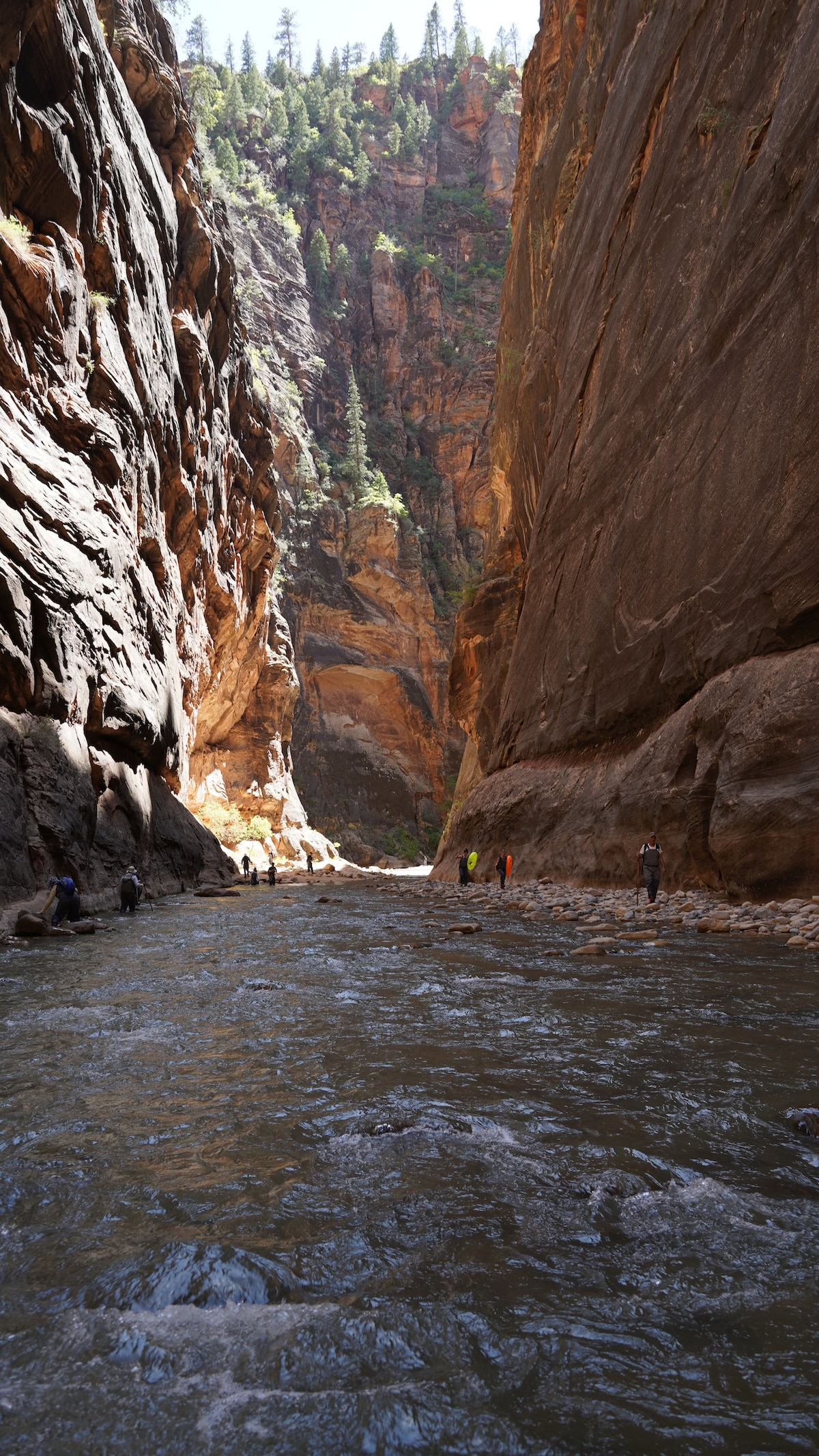 A scenic view of a narrow canyon reveals towering rock walls, showcasing rich textures and colors. A gentle river flows through the center, with individuals engaged in various activities along the water's edge. Sunlight filters through the canyon, highlighting the contours of the landscape.