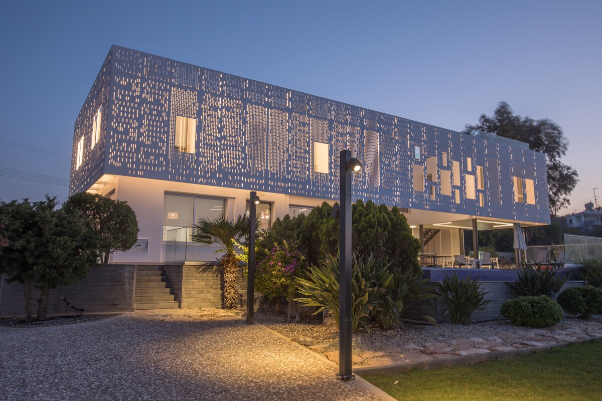 The modern villa features a striking façade with a unique pattern illuminated by evening lights. Surrounded by landscaped gardens, the entrance is approached via decorative stone pathways, leading to an inviting outdoor space with seating and glimpses of the pool area in the background.