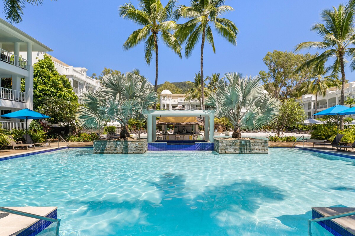 A spacious swimming pool glistens under clear blue skies, surrounded by landscaped tropical plants and palm trees. A pool bar is situated at the end of the pool, and shaded lounge chairs invite relaxation along the water’s edge.