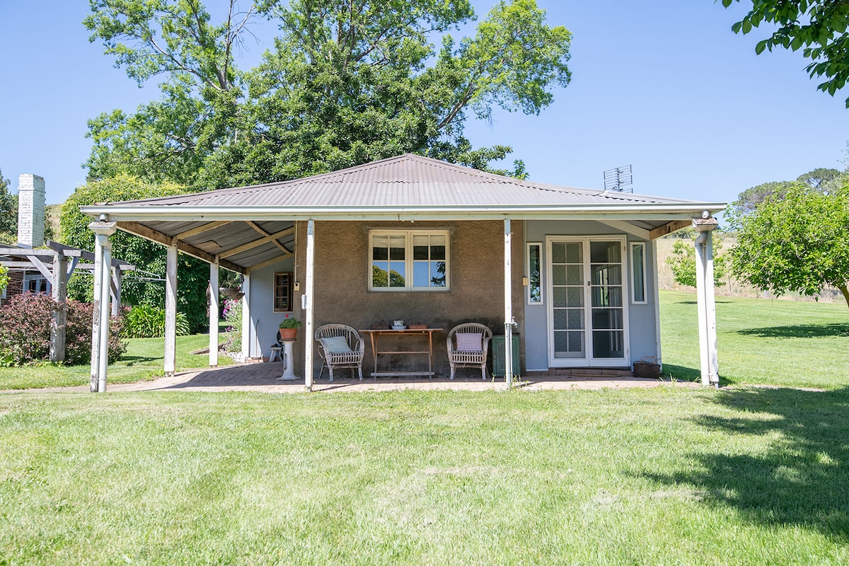 A charming cottage is set on a grassy area, surrounded by trees and open fields. A covered porch features two outdoor chairs and a small table, inviting relaxation. Large windows allow natural light to illuminate the cozy interior while providing views of the surrounding garden.