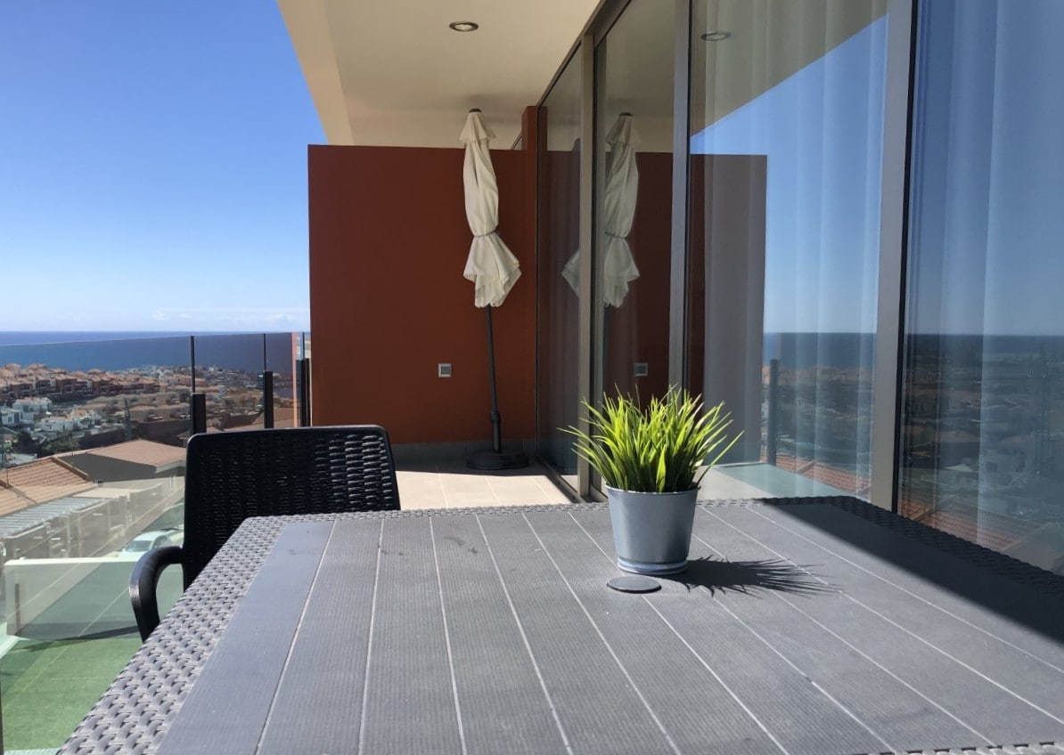 A private balcony features a sleek table with a potted plant at its center. Glass doors reflect the coastal view and sunlight, enhancing the sense of openness. The clear blue sky and distant ocean can be seen in the background, contributing to a calm atmosphere.