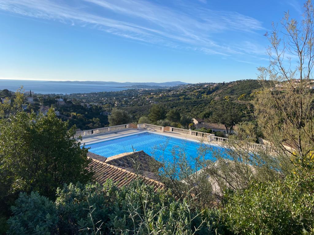The image captures a panoramic view of a large swimming pool surrounded by greenery, overlooking rolling hills leading to the sea. The bright blue water reflects the clear sky, while the distant coastline and mountains provide a serene backdrop.