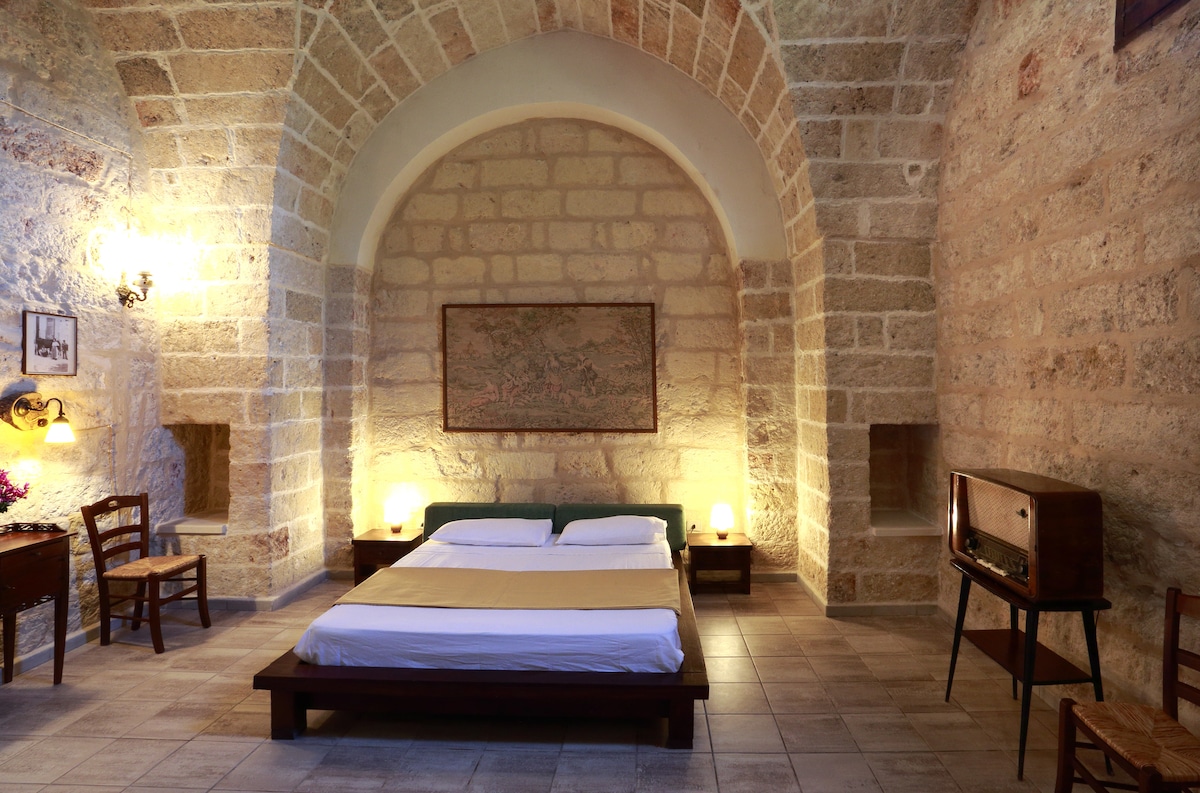 A spacious room features high arched ceilings with exposed stone walls, creating a rustic atmosphere. A large bed occupies the center, flanked by bedside lamps. A vintage radio sits on a wooden table, accompanied by a simple wooden chair. Artwork adorns the walls.