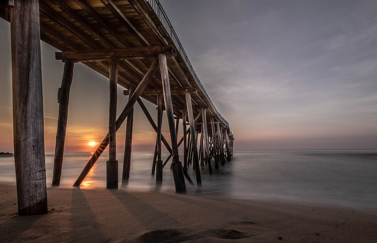 A wooden pier stretches over the calm water, supported by sturdy columns. The sun sets in the background, casting a warm glow on the sandy shore. Gentle waves lap against the pilings, creating a peaceful coastal scene.