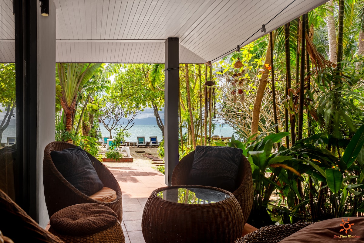 A shaded outdoor seating area features comfortable wicker chairs arranged around a round glass table. Lush tropical plants surround the space, with views of the beach and sea visible in the background.