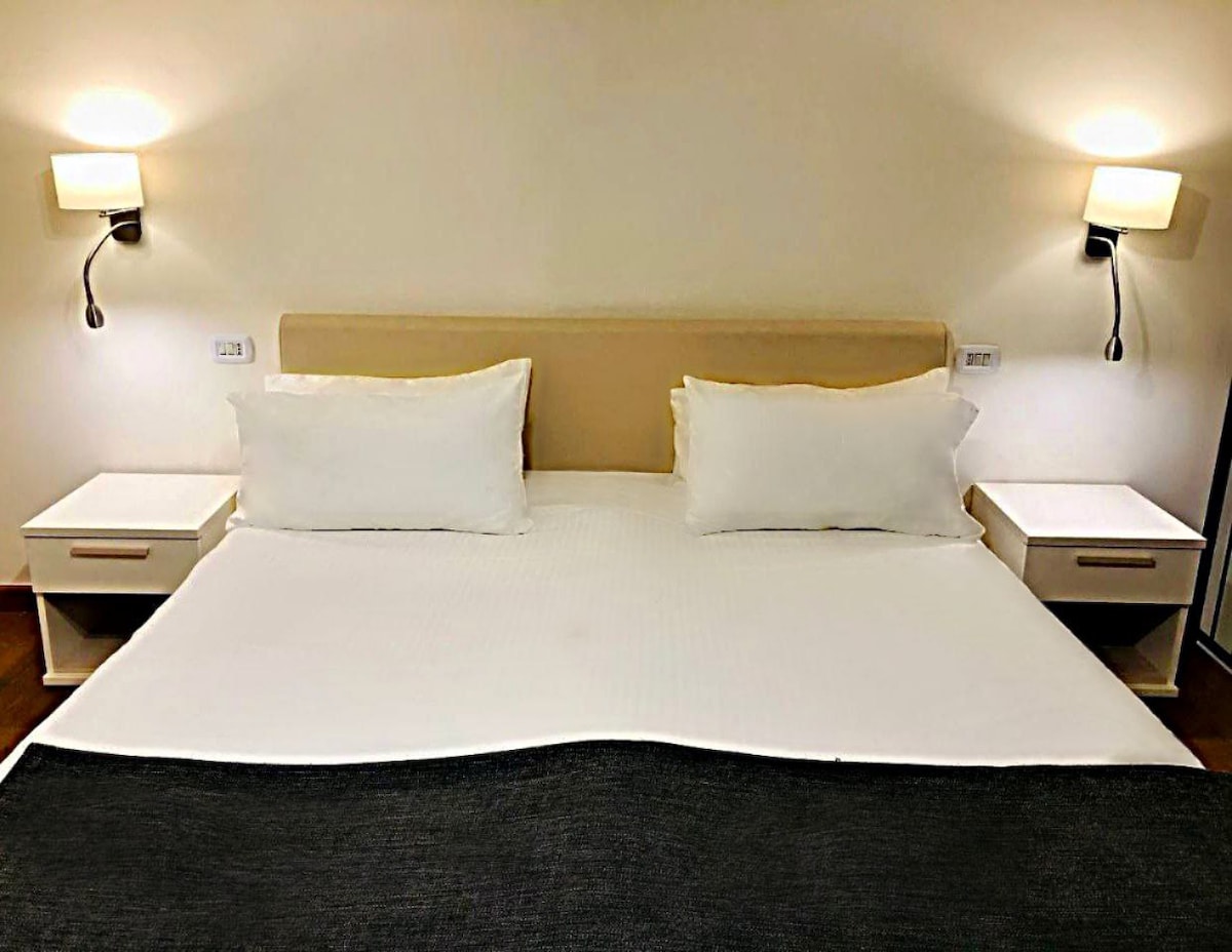 A spacious bed with crisp white linens is flanked by two bedside tables, each featuring a simple lamp. Soft lighting creates a calm atmosphere, while the dark textured blanket is neatly draped at the foot of the bed. Minimalist decor emphasizes the serene setting.